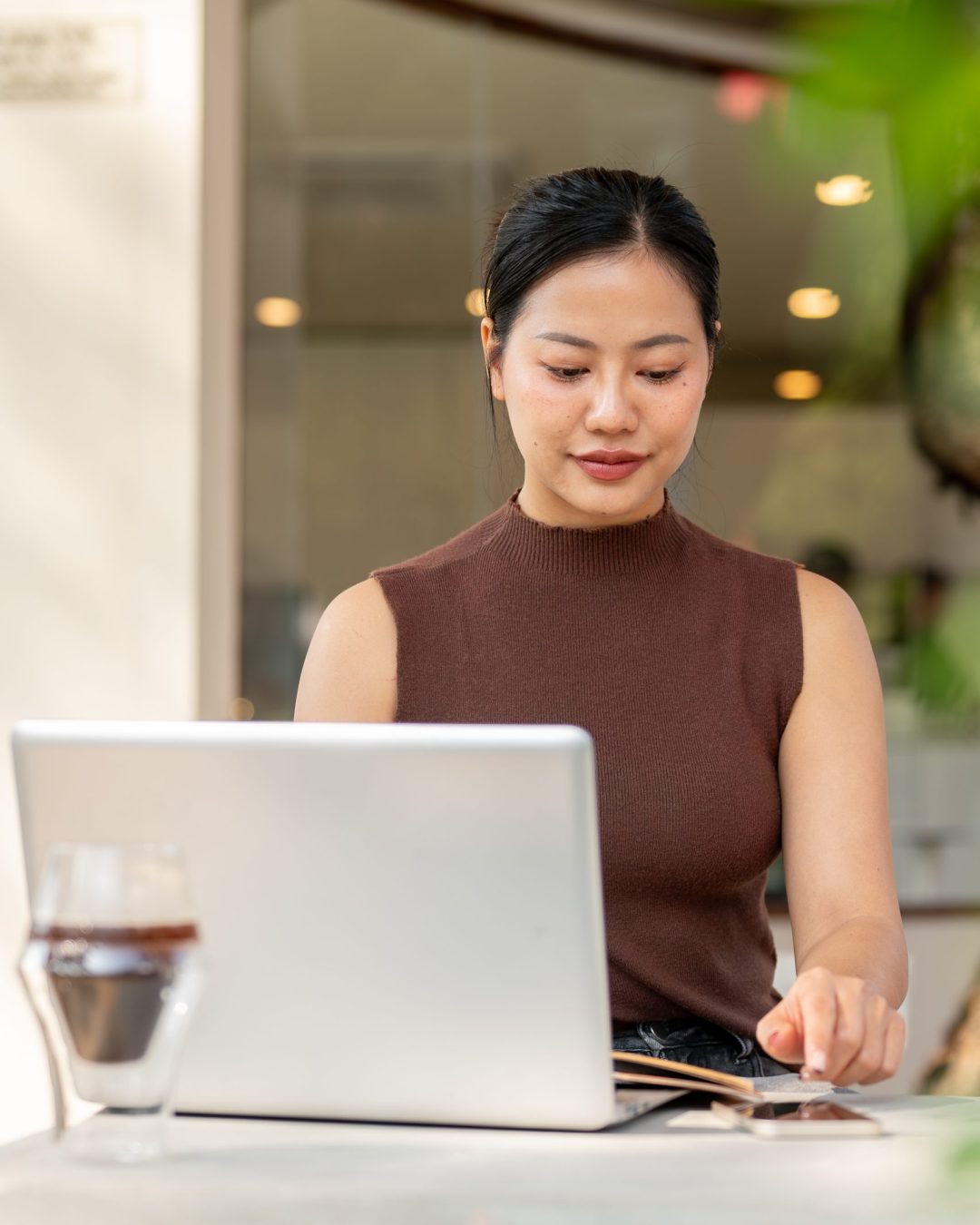 A beautiful, satisfied Asian woman checks messages on her smartphone while working remotely from a coffee shop, seated in front of a laptop.